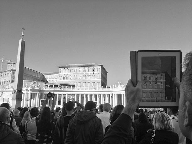 Vatican City, Vatican - November 2, 2014: a usual crowd gathers in front of the Pope at midday on Sundays. The Pope is visible out of his balcony in the image captured by a man on his iPad's screen. Other people use digital devices to capture images of the event. 