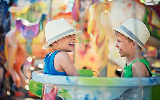 Two little brothers sitting in the same cup on carousel. The boys are aged 5 and wearing fedoras. Boys are having fun and laughing.