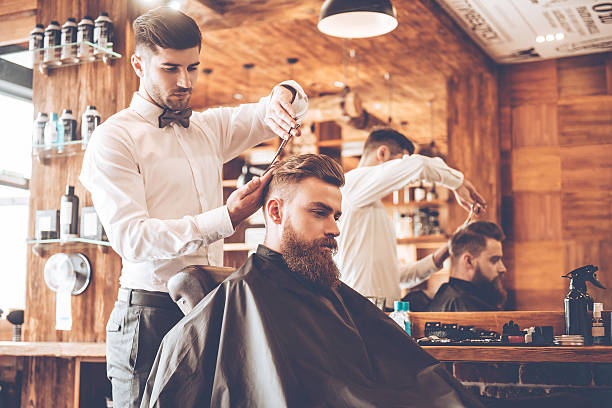 Everything should be perfect. Side view of young bearded man getting haircut by hairdresser while sitting in chair at barbershop barber shop stock pictures, royalty-free photos & images