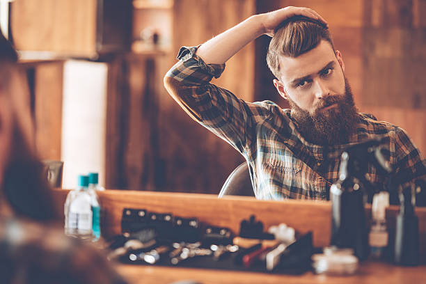 Time for new haircut. Handsome young bearded man looking at his reflection in the mirror and keeping hand in hair while sitting in chair at barbershop men hair cut stock pictures, royalty-free photos & images