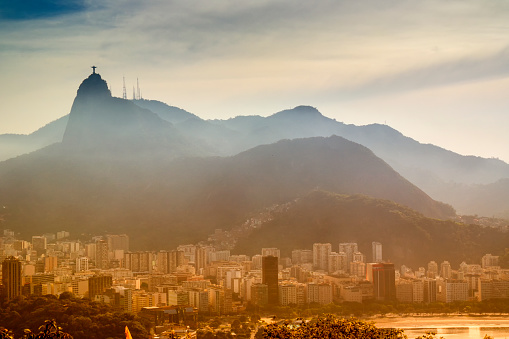 Aerial view of Rio de Janeiro at sunset.  The beach is visible in the bottom-right section of the photo.There is a small forested area in the middle of the photo.  Buildings surround the beach and forest area, extending toward the mountains in the background.  The mountains are blanketed by mist that partially obscures them, but the Christ the Redeemer statue is visible above the mist.  The sunlight adds a warm glow to the photo, and the area behind the mountains is golden.  