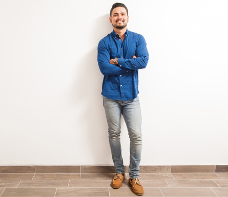 Full length portrait of a young and confident Hispanic man standing against a white wall with his arms crossed and smiling