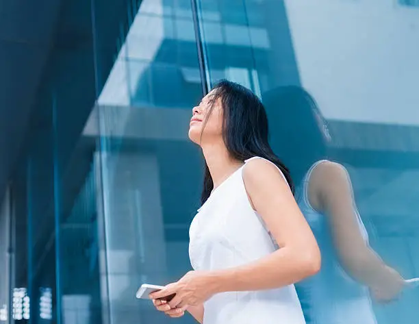 asian businesswoman standing against the glass wall with holding cellphone in hand outside of the office building.