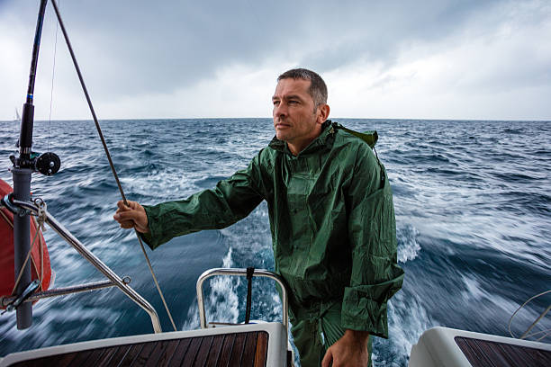 Young skipper of sailboat yacht standing on ship deck in the green rain suit. Boat is sailing very fast due to trail in water.