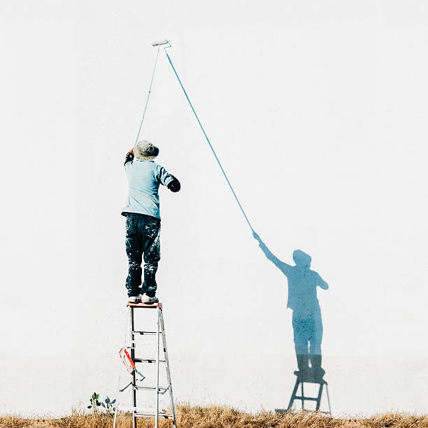 Home Improvements Hispanic man on top of a ladder painting a large facade on white with a paint roller. house painter ladder paint men stock pictures, royalty-free photos & images