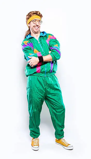 A cool, funky young adult in late 1980's / early 1990's fashion style, with mullet, fluorescent colored track suit, nerdy glasses, and sweat band.   He smiles with a cheesy grin, his arms folded.  Vertical full body image, isolated on a white background.