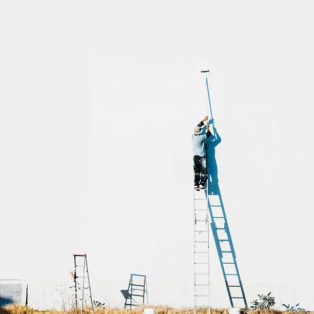 Hispanic man on top of a ladder painting a large facade on white with a paint roller.