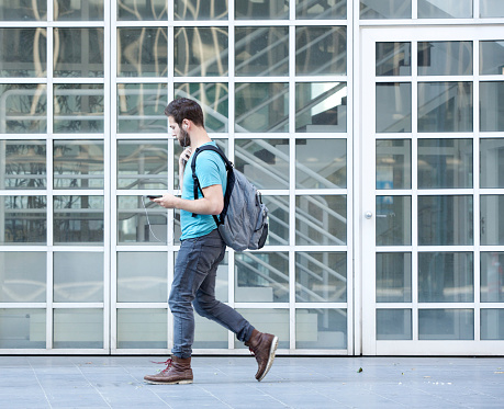 Side view portrait of a male student walking on campus with bag and mobile phone