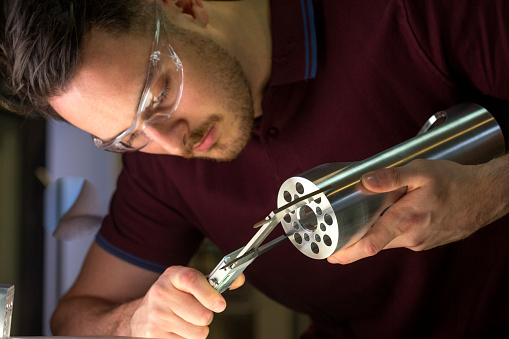 Male using a vernier device to measure and inspect a metal object he is holding. The man is wearing safety goggles. Close up