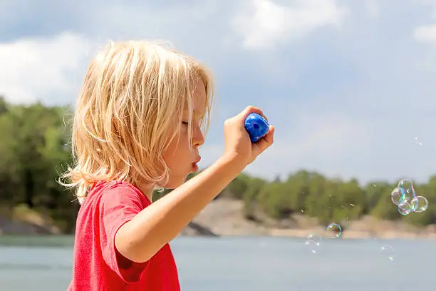 6 years old blond longhaired boy in red T-shirt blowing soap bubbles. Blurred background Stockholm Archipelago, Sweden.