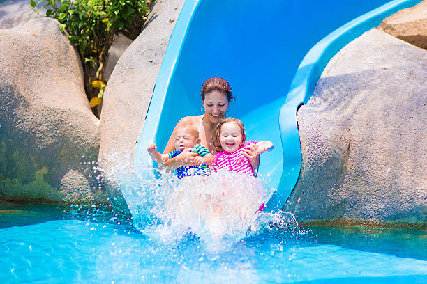 Young happy mother and two children, baby boy and toddler girl having fun at water slide in a tropical pool on a hot summer day