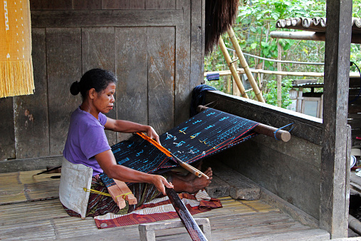 Bena, Indonesia - August 17, 2014: A woman weaving textiles at the traditional hill tribe village of Bena, home to the Ngada ethnic group, located in the mountains of central Flores. Much of the layout and structures are as they have been for hundreds of years.