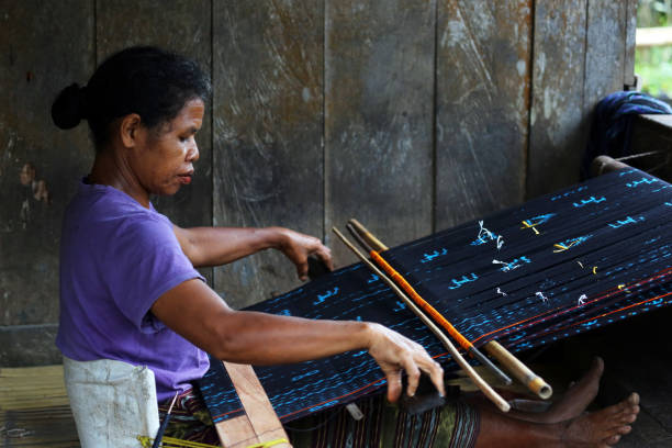 Bena, Indonesia - August 17, 2014: A woman weaving textiles at the traditional hill tribe village of Bena, home to the Ngada ethnic group, located in the mountains of central Flores. Much of the layout and structures are as they have been for hundreds of years.