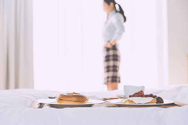 asian businesswoman standing by window, breakfast on the bed  in the morning at  hotel, business trip on the road. Focus on the food.