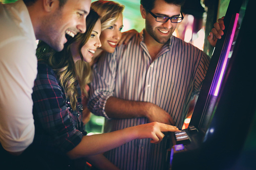 Group of young adults in late 20's playing slot poker and fruits and wining.Wearing casual clothes and having fun on weekend night. There are many slot machines out of focus in background.
