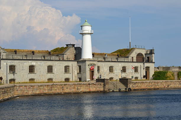 A coastal navigation beacon painted red and white rises above a rocky breakwater, standing against the backdrop of a calm lake and partly cloudy sky. The peaceful setting evokes themes of maritime guidance, security, and nature.