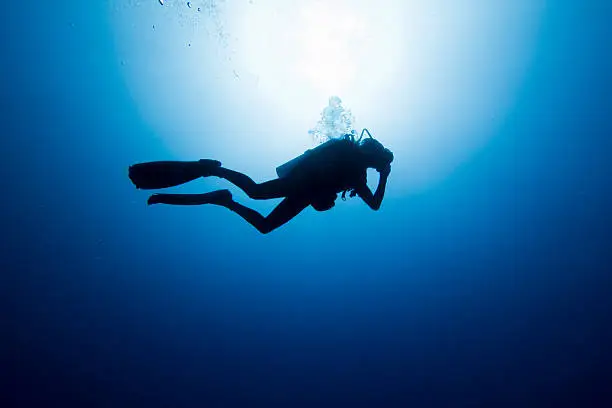A DSLR photo of a diver deep in Tiputa Pass, Rangiroa, French Polynesia. The camera is in a low angle view and the diver is silhouetted against a bright sunburst in the surface. From the sunburst to the bottom of the image there is a gradient from white to black passing through all shades of blue.