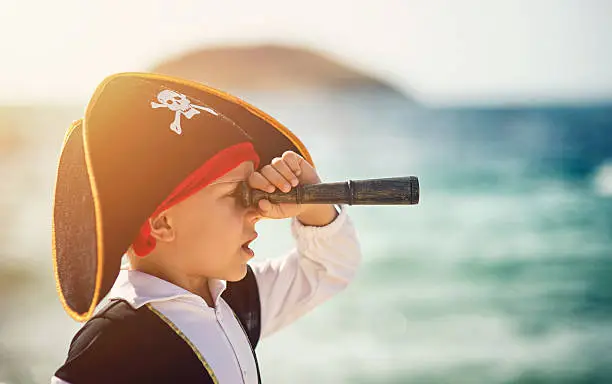 Little boy dressed up as pirate looking through spyglass. The boy is aged 5 and he is very suprised. Sunny summer day.