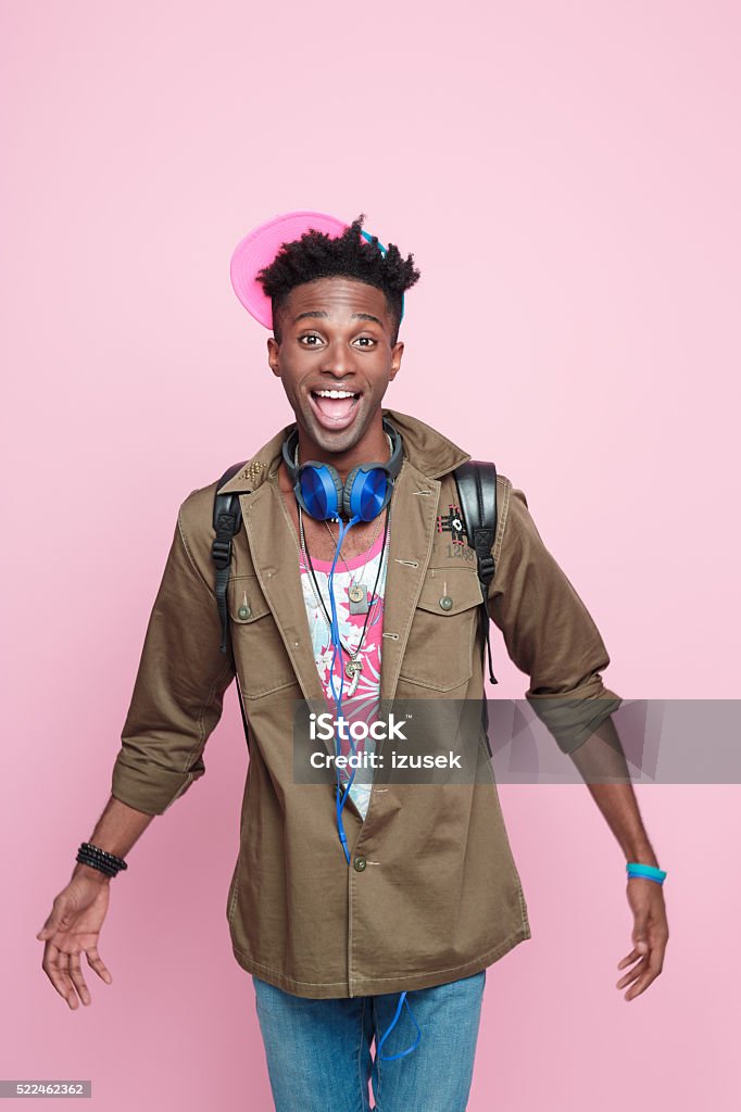 Studio portrait of funky, excited afro american young man Summer portrait of happy, cool afro american young man in modern outfit, wearing headphone, cap, jacket and backpack, standing against pink background, laughing at camera. African Ethnicity Stock Photo