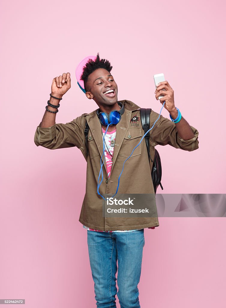 Studio portrait of cool, excited afro american young man Summer portrait of happy, funky afro american young man in modern outfit, wearing headphone, cap, jacket and backpack, standing against pink background, holding a smart phone in hand. Men Stock Photo