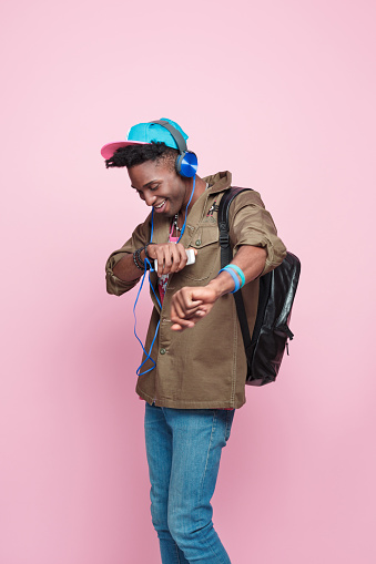Summer portrait of happy, cool afro american young man in modern outfit, wearing headphone, cap, jacket and backpack, dancing against pink background.