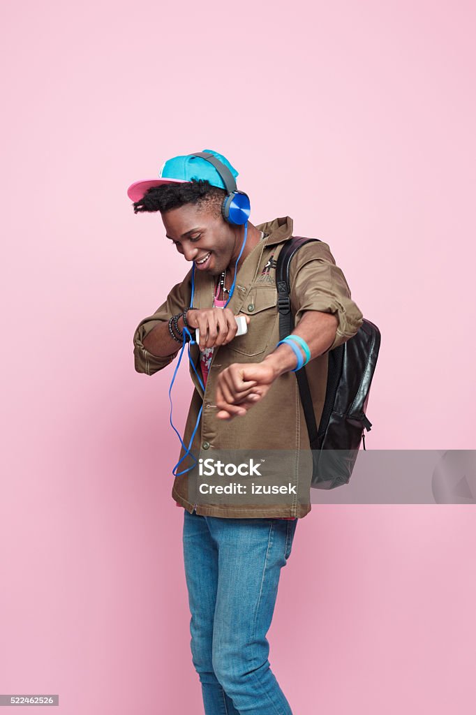 Studio portrait of funky, excited afro american young man Summer portrait of happy, cool afro american young man in modern outfit, wearing headphone, cap, jacket and backpack, dancing against pink background. Men Stock Photo