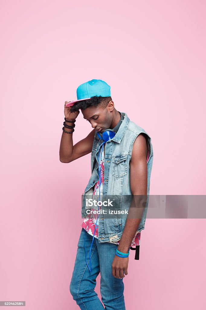 Summer portrait of funky afro american guy Fashion portrait of afro american young man wearing headphone, cap and jeans sleeveless jacket, standing against pink background. Adult Stock Photo