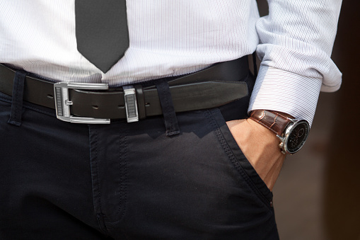 Closeup shot of male waist with hand in pocket dressed in black pants, belt, grey shirt, black tie and watch with brown watch strap