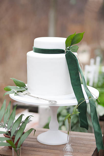 White cake decorated with green velvet ribbons and leaves with silver cake shovel on white stand and geen and beige background