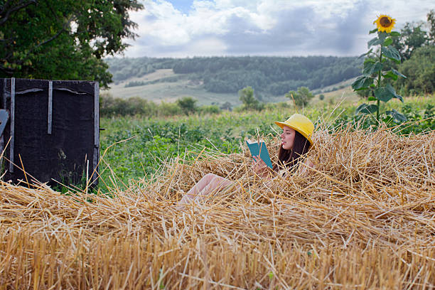 Village. Young girl in a straw hat sitting on straw and reading a book. Right growing sunflower. On the left is the old well.
