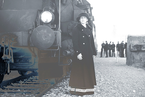 Attractive woman in retro styled coat, dress and hat posing at the steam train - 1910s style. Black and white blue toned image.