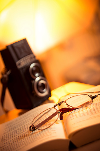 Old mans glasses on an opened book on the table. There is a film camera on background.