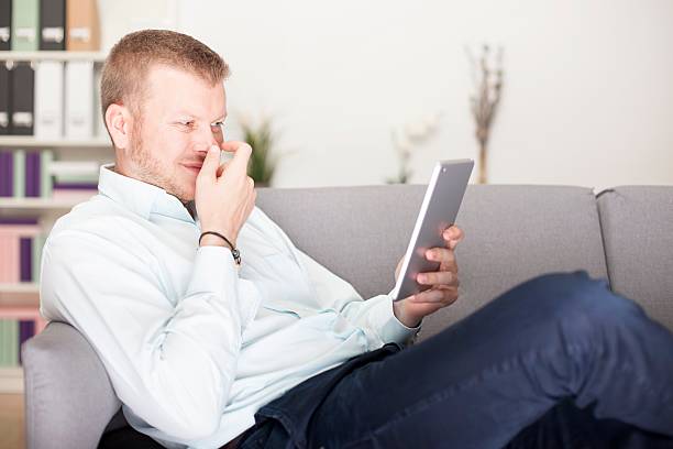 Man snickering to himself as he reads his tablet computer holding his hand in front of his face as he lies back comfortably on a sofa at home