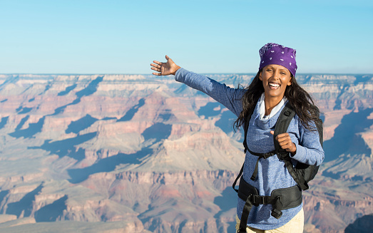Smiling young backpacker woman showing with excitement the Grand Canyon scenic view, looking over the camera.