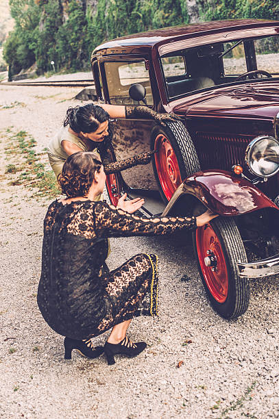 Two women in festive clothings trying to change the wheel of a car - 1939 style. Desatured and toned image.