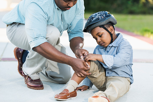 An African American father putting an adhesive bandage on the knee of his 5 year old boy. His son must have fallen off his bike or scooter since he is wearing a helmet.
