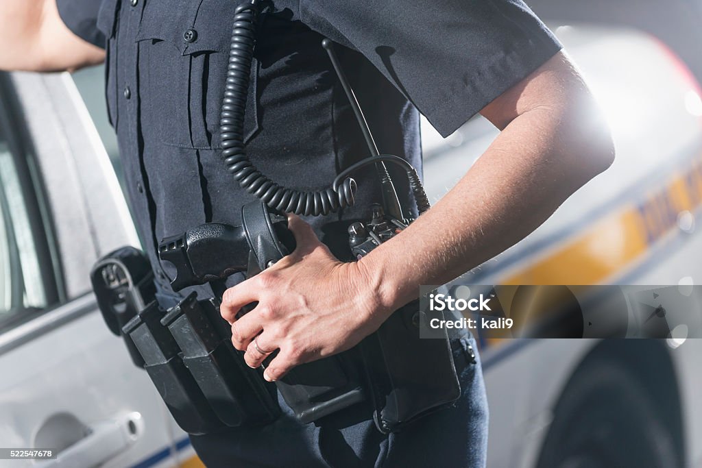 Cropped view of policewoman next to patrol car A cropped view of a female police officer standing next to her police car. Her hand is resting on her tool belt with gun holster and radio. Police Force Stock Photo