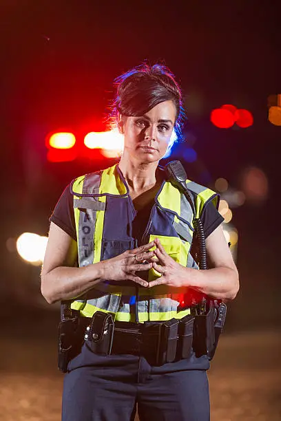 A policewoman wearing a safety vest standing in the street at night. In the background, her police car is out of focus, with lights illuminated. She has a serious expression on her face.
