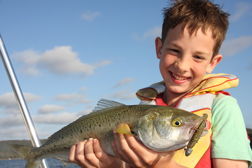 A digital SLR photograph of a smiling boy holding a large Australian salmon.  The boy is wearing a colorful life jacket.  The blue sky can be seen in the background. The photo was taken at Mallacoota in Victoria, Australia.