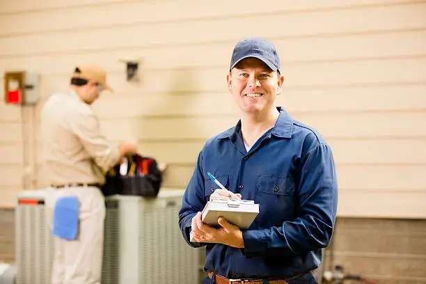 Caucasian and Latin descent repairmen work on a home's air conditioner unit outdoors. One foreground is writing on clipboard. Other man in backgroud is checking unit using the tools in his tool bag.  They both wear uniforms. 