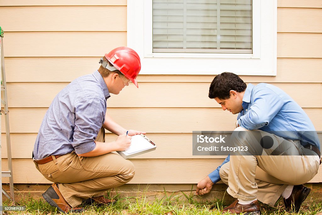 Inspectors or blue collar workers examine building wall, foundation. Outdoors. Repairmen, building inspectors, exterminators, engineers, insurance adjusters, or other blue collar workers examine a building/home's exterior wall and foundation. One wears a red hard hat and clear safety glasses and holds a clipboard.  The other checks the foundation with tool.  Examining Stock Photo