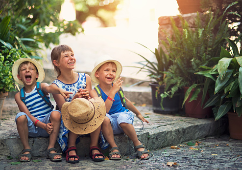 Three kids tourist resting in the beautiful mediterranean town. Little boys are wearing backpacks and hats. Theirs sister is wearing sundress. Kids are sitting on stone stairs and laughing.