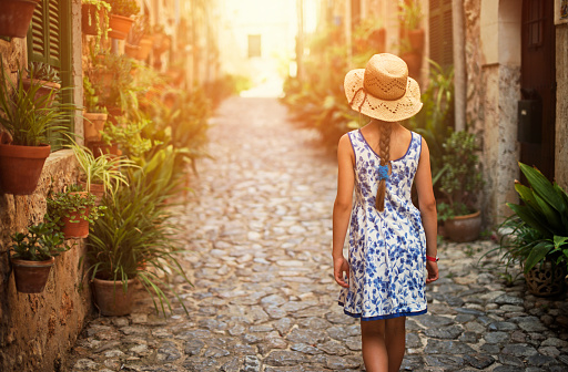 Little girl walking down beautiful street of a mediterranean town. The girl is wearing a sundress and straw hat.