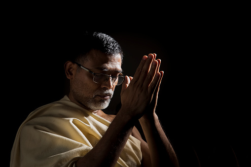 An old Hindu man praying. His head is bowed in reverence and hands are folded. He is wearing glasses. This is a customary way of praying among followers of Hinduism. Shot in studio with black background.