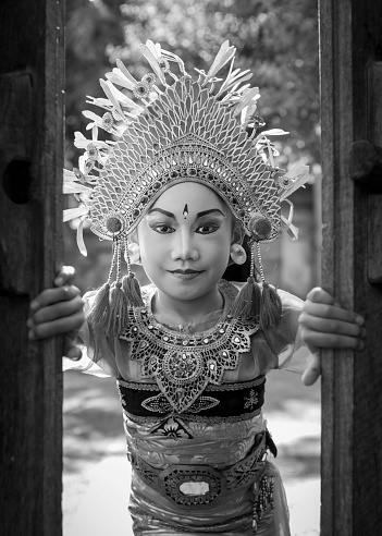 A 6 year old cute Asian little girl wears a traditional Bali costume and stares through a wooden carved door in a temple location. Children at a very early age learn the ancient art of Ramayana dancing and other Hindu dances generally located in temple schools. In a Black and White mode to underline the ancient culture of Bali.