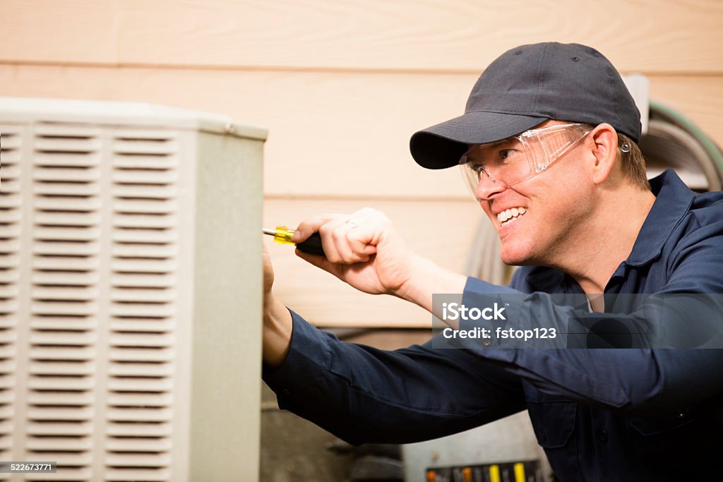 Air conditioner repairman works on home unit. Blue collar worker. Repairman works on a home's air conditioner unit outdoors. He is checking the compressor inside the unit using a screwdriver. He wears a navy blue uniform and his safety glasses.  Close up. Air Conditioner Stock Photo