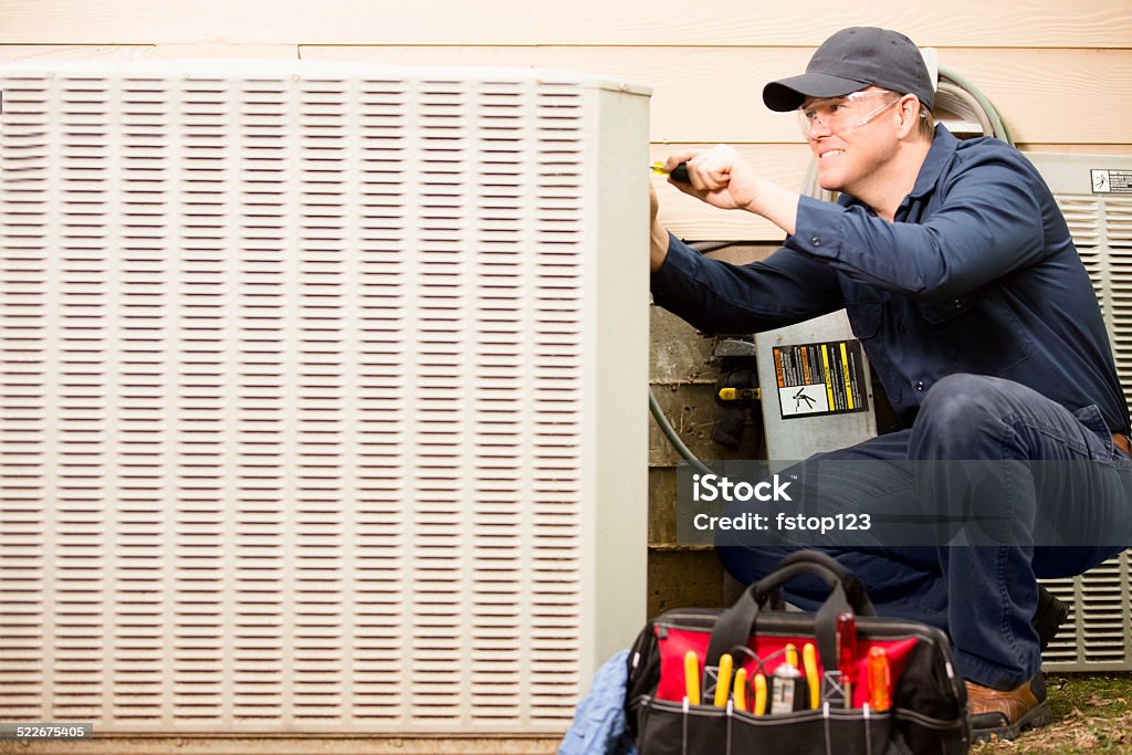 Air conditioner repairman works on home unit. Blue collar worker. Repairmen works on a home's air conditioner unit outdoors. He is checking the compressor inside the unit. He has his tools and tool bag beside him. He wears a navy blue uniform and his safety glasses.  Air Conditioner Stock Photo