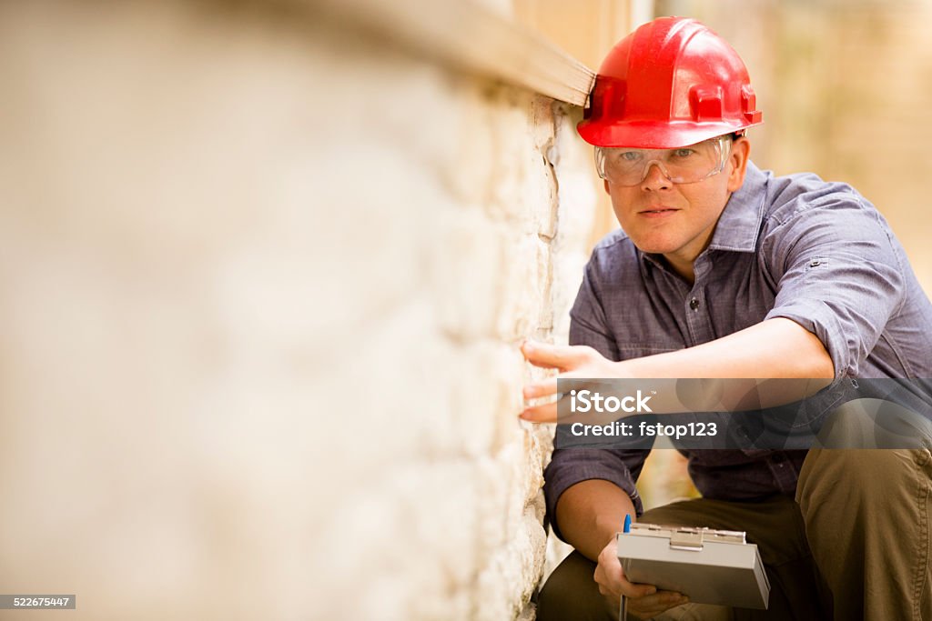 Inspector or blue collar worker examines building wall outdoors. Repairman, building inspector, exterminator, engineer, insurance adjuster, or other blue collar worker examines a building/home exterior wall.  He wears a red hard hat and clear safety glasses and holds a clipboard.  Examining Stock Photo