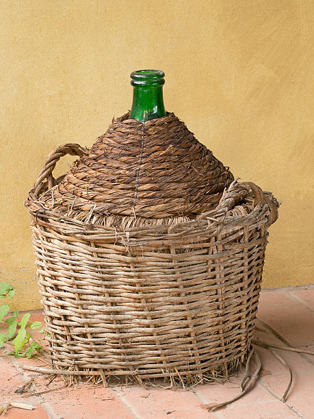 istock Antique old demijohn, carboy, by yellow wall. Wicker basket, 522714629