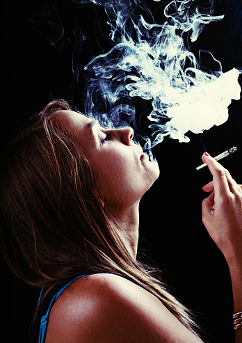 Profile of a young woman, probably stoned, puffing out smoke after taking a toke on a joint of marijuana. A quiet rebel. Moody black background with exhaled smoke..